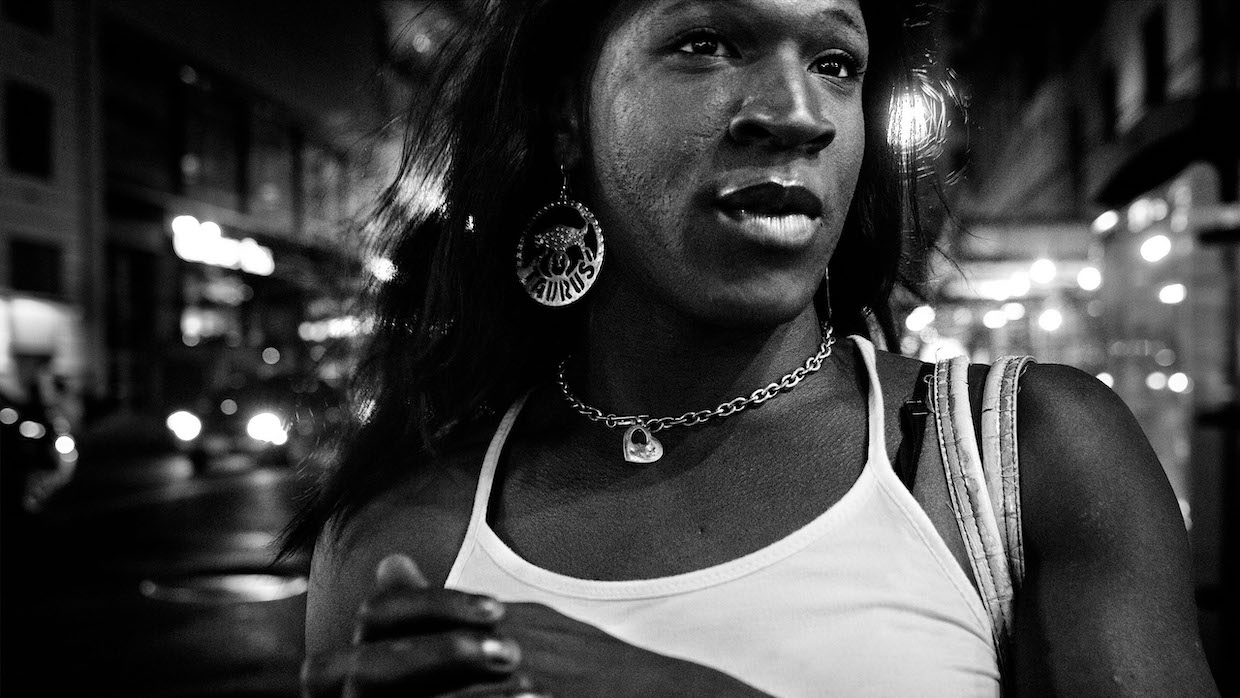 A Black trans woman wears a white tank top, a chain necklace and large hoop earrings that say "Taurus" with a picture of a bull. She walks in NYC's meatpacking district behind her.