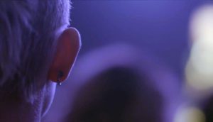 Someone's right ear featuring a single black horseshoe-shaped hoop. They are bathed in indigo-colored lighting, likely at a music venue.