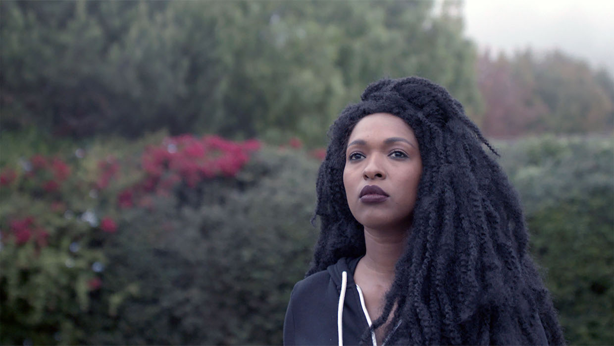 A woman with locs and dark purple lipstick stands in front of flowers and shrubbery.