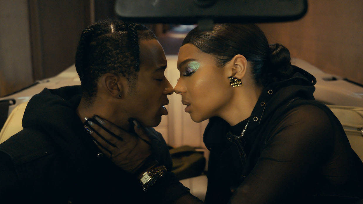 Two people lean in for a kiss, one wearing metallic eyeshadow. They are both dressed in all-black.