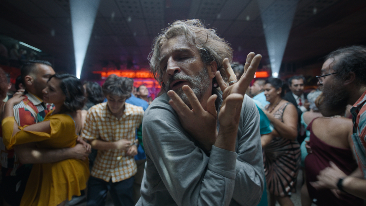 A man with shoulder-length gray hair dances in a crowded club.