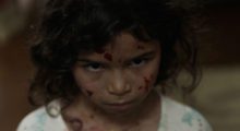 A young girl with curly brown hair stands with her head tilted downward, her face, neck and shirt dotted with streaks of blood.