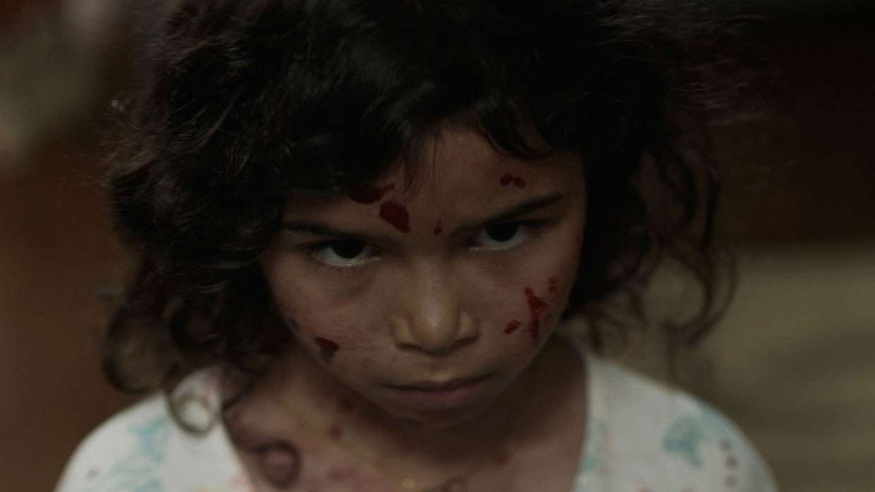 A young girl with curly brown hair stands with her head tilted downward, her face, neck and shirt dotted with streaks of blood.