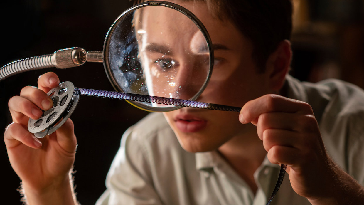 A boy wearing a tan polo shirt examines a strip of film under a magnifying glass.