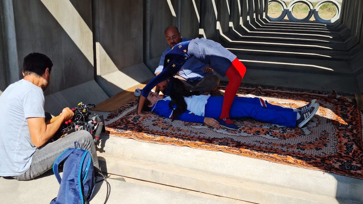 A cinematographer crouches down to get a shot of a woman bending backwards over a man, both wearing bright blue outfits.