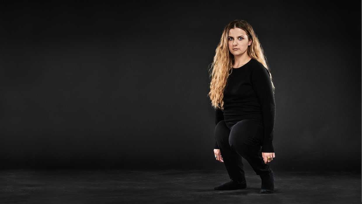 A woman with long, curly blond hair wears all black and stands to the right amid a gray backdrop.