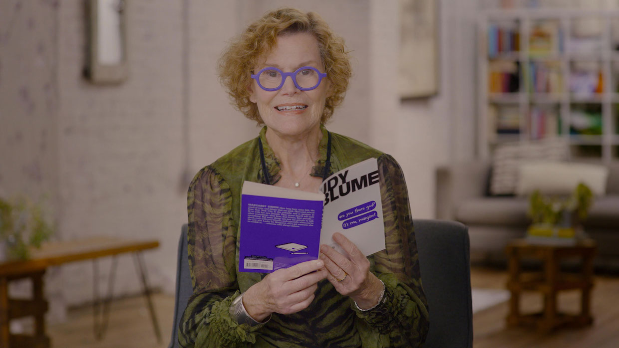 A woman with short, curly red hair sits in a chair with an open copy of Are You There, God? It's Me, Margaret.