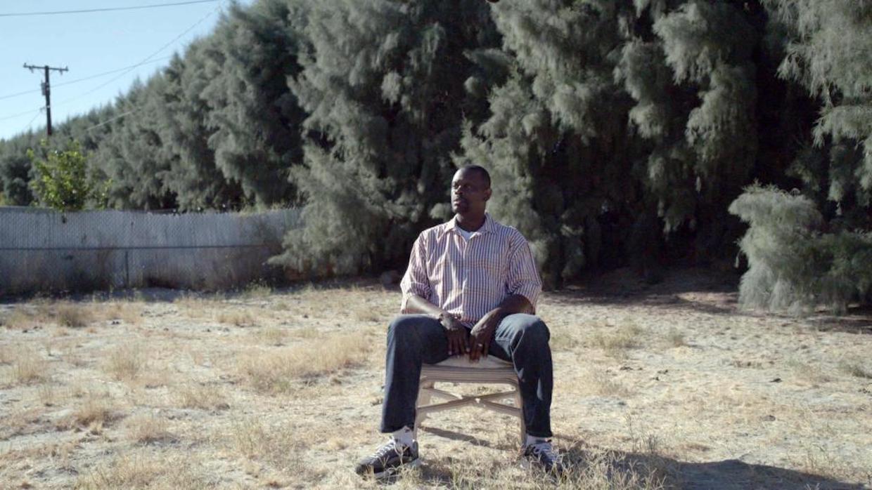 A Black man sits in a chair outdoors, with dead grass covering the ground and an imposing line of thick trees cutting through the background.