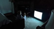 Two children sit in front of a glowing '90s-era TV set on a carpeted floor in the ominous darkness of their living room.