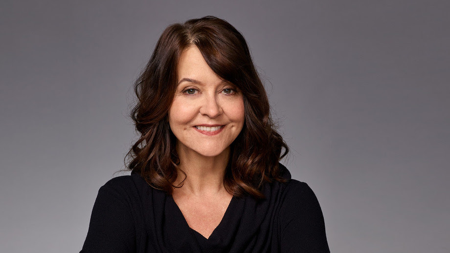 A woman with brown hair and a black scoop next top smiling against a grey backdrop