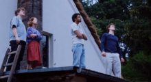 Four young people stand on top of a roof, gazing at something unseen in the dusky sky.