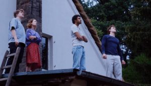 Four young people stand on top of a roof, gazing at something unseen in the dusky sky.