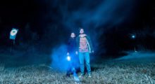 A teenage boy and girl stand in an open field with a flashlight as a mist rolls in.