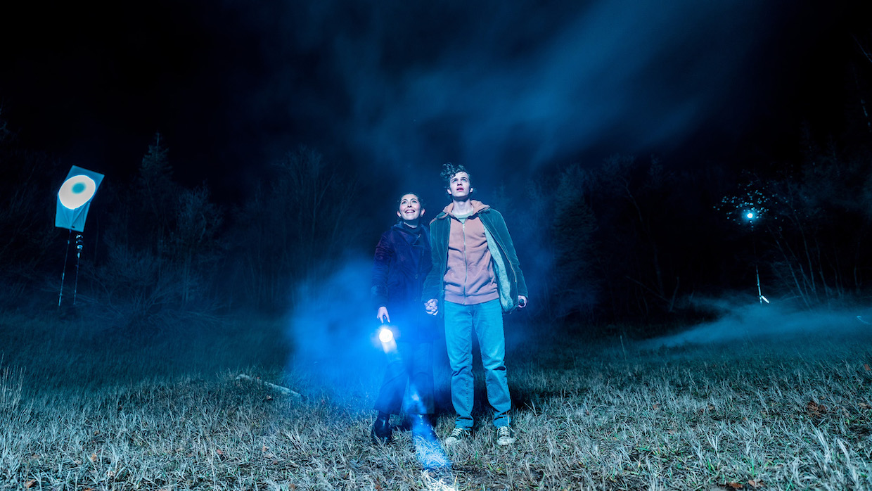 A teenage boy and girl stand in an open field with a flashlight as a mist rolls in.