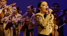 A mariachi band performs on stage, with a woman violinist singing into a microphone.