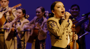 A mariachi band performs on stage, with a woman violinist singing into a microphone.