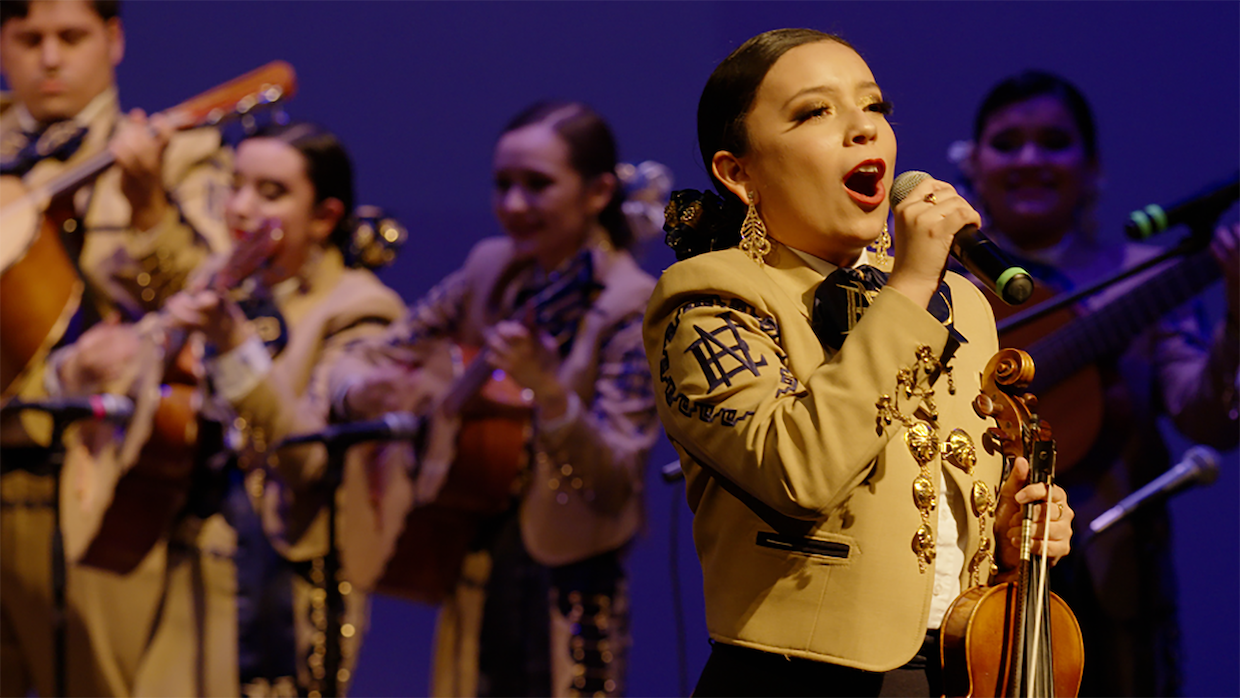 A mariachi band performs on stage, with a woman violinist singing into a microphone.