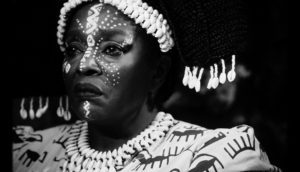 An African woman frowns, she dons elaborate face paint and shells woven into her hair. She is shot via black and white photography.