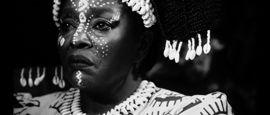 An African woman frowns, she dons elaborate face paint and shells woven into her hair. She is shot via black and white photography.