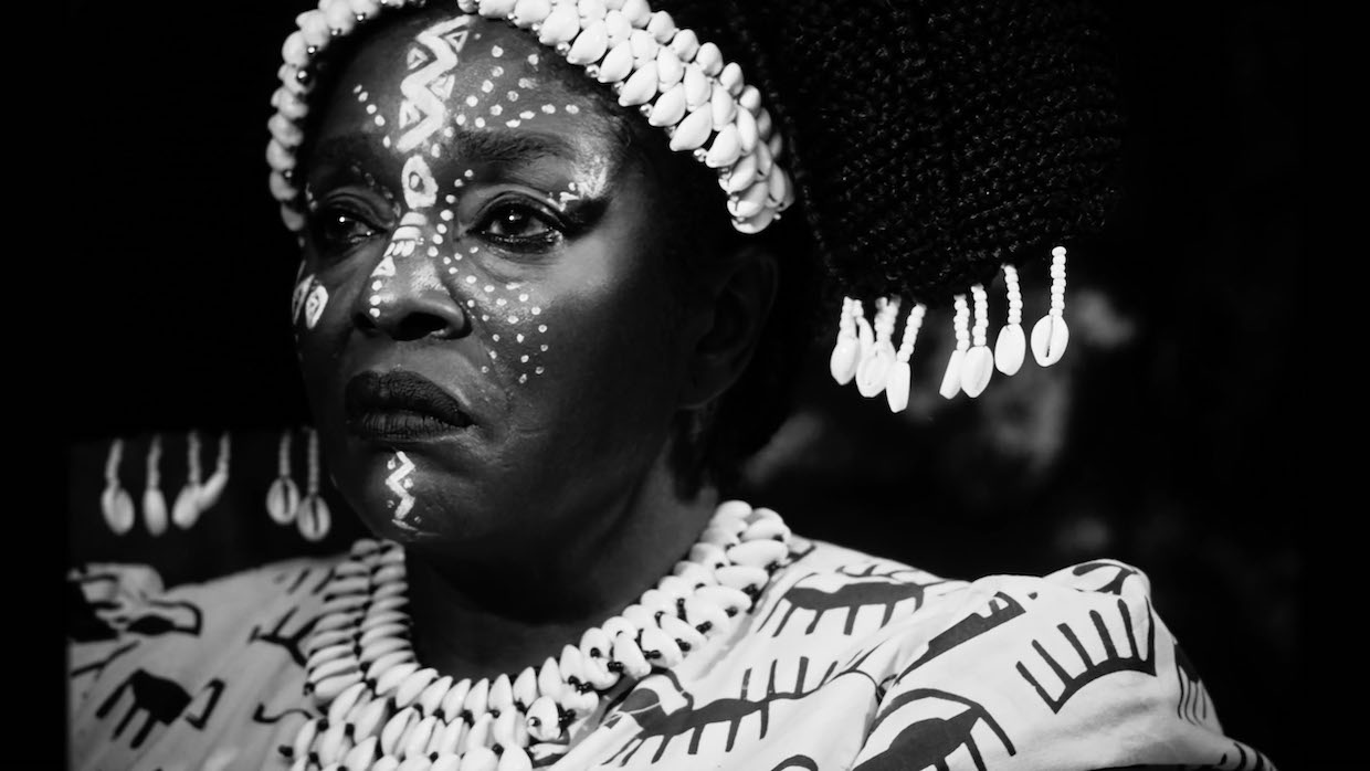 An African woman frowns, she dons elaborate face paint and shells woven into her hair. She is shot via black and white photography.