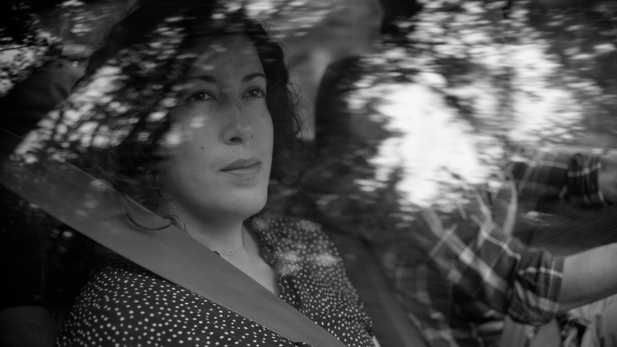 A black and white image of a woman sitting in the passenger seat of a car looking out the window. She has shoulder-length wavy hair and wears a blouse with small polka dots on it.