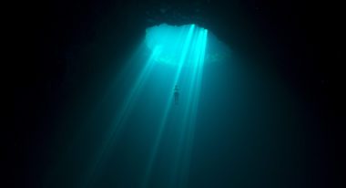 A freediver ascends through an undersea cave as a bright blue sunbeam shines down on them.