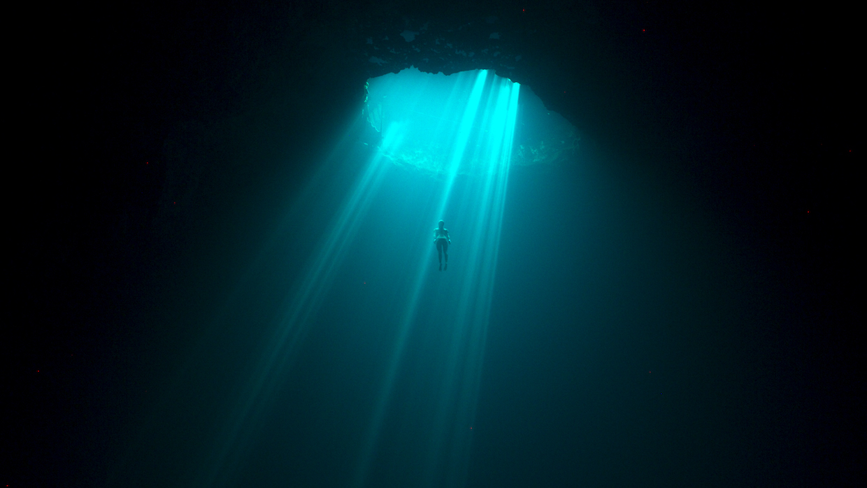 A freediver ascends through an undersea cave as a bright blue sunbeam shines down on them.