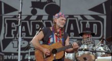 Willie Nelson performs on stage with an acoustic guitar. He dons a red, white and blue bandana and guitar strap and his distinctive braids.