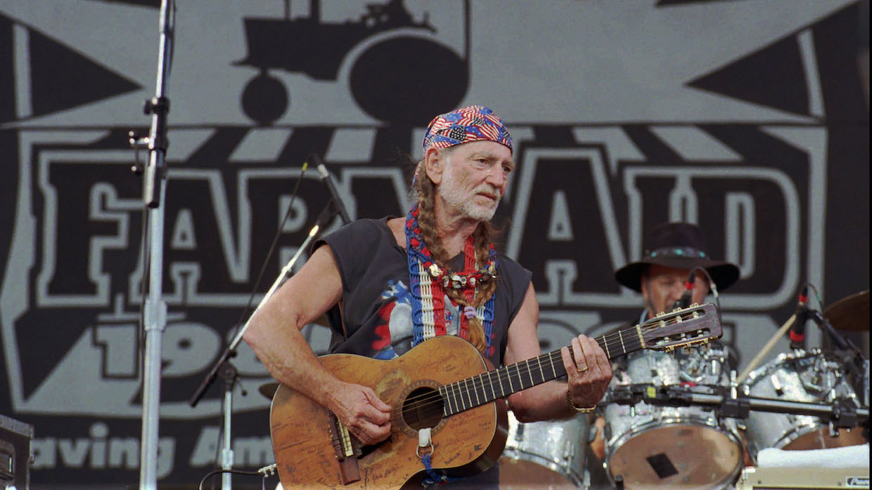 Willie Nelson performs on stage with an acoustic guitar. He dons a red, white and blue bandana and guitar strap and his distinctive braids.