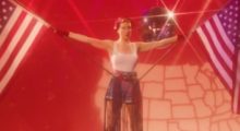 A woman wearing a white tank top and denim shorts holds two American flags, one in each hand, with her arms outstretched at her side. A disco ball and map of the U.S. are behind her.