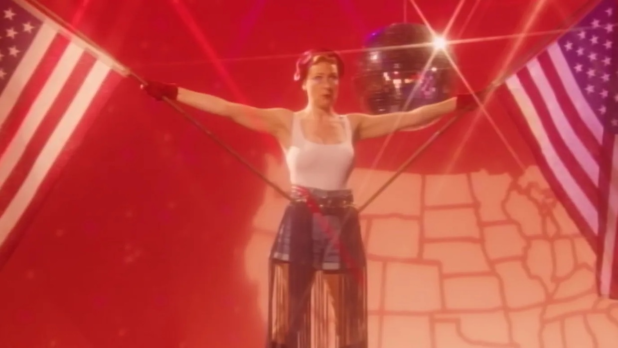 A woman wearing a white tank top and denim shorts holds two American flags, one in each hand, with her arms outstretched at her side. A disco ball and map of the U.S. are behind her.