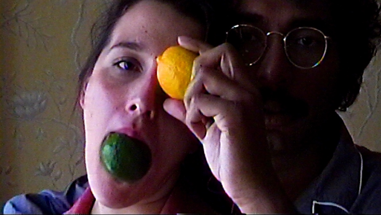 A man and a woman stand next to each other. The woman holds a lime in her mouth and places a lemon over her left eye. The man wears glasses and his face is semi-obscured by her head and arm.