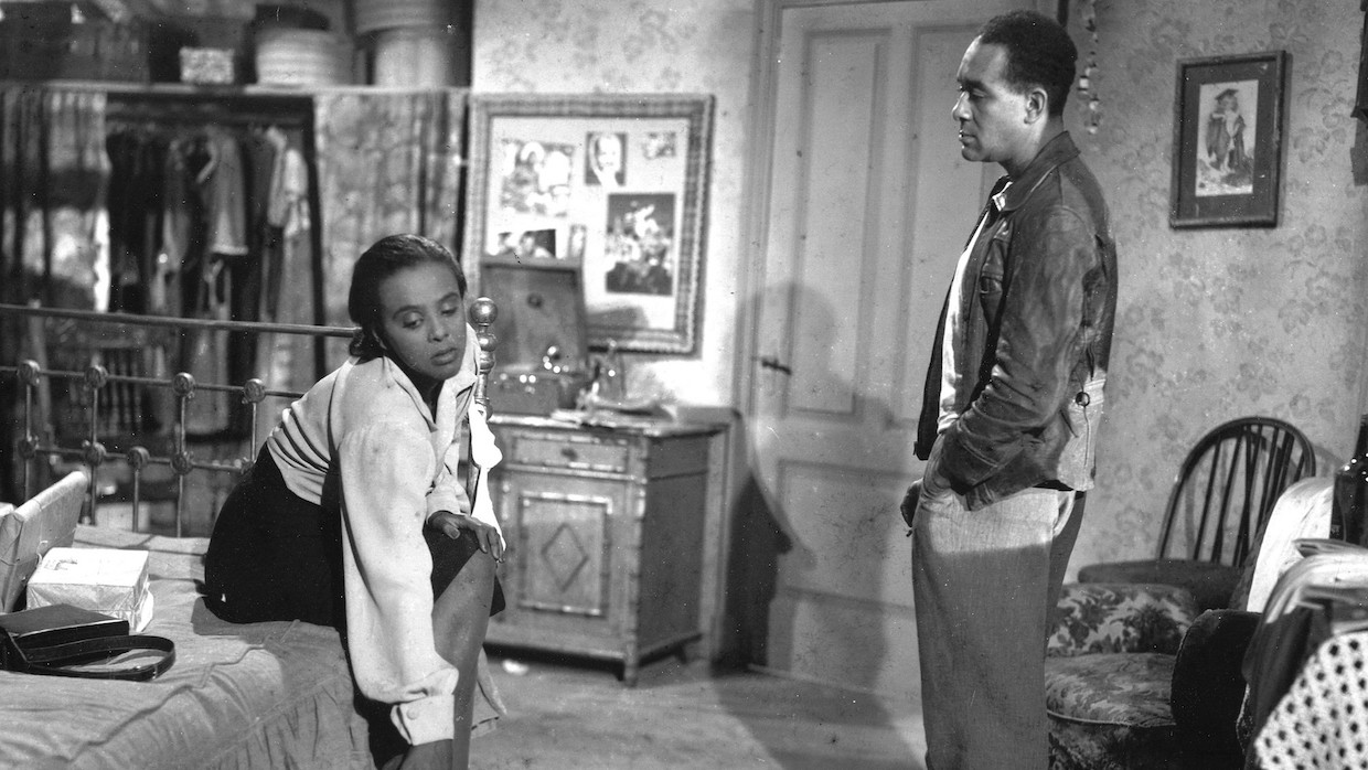 A Black man and woman are in a room together. The woman sits on the bed looking away from the camera while the man stands a few feet away and looks straight at her.
