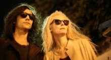 A man with shoulder length black hair and a woman with long platinum hair walk side by side with sunglasses on in the dead of night.
