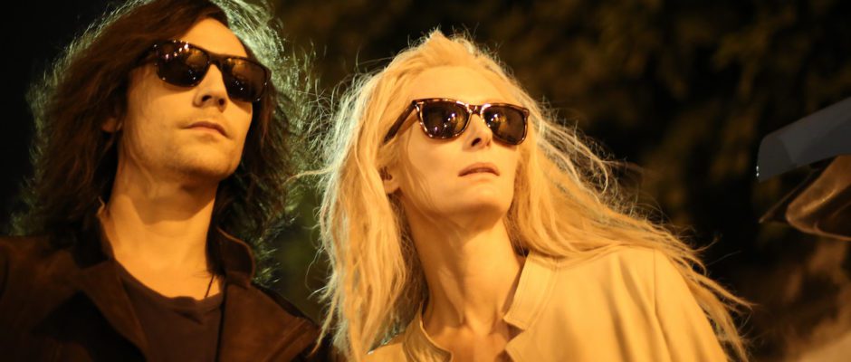 A man with shoulder length black hair and a woman with long platinum hair walk side by side with sunglasses on in the dead of night.