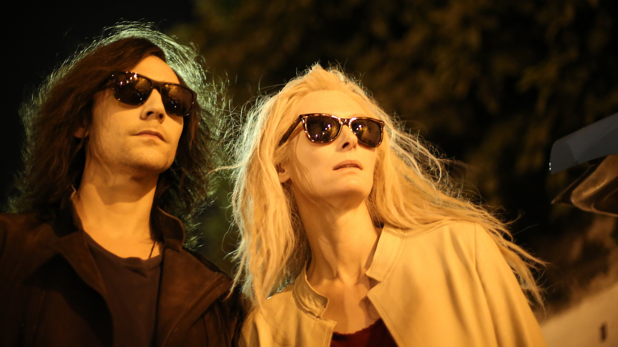 A man with shoulder length black hair and a woman with long platinum hair walk side by side with sunglasses on in the dead of night.