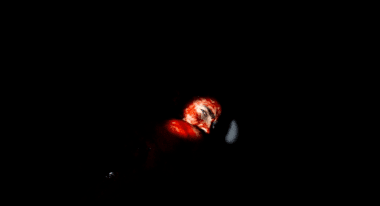 In the pitch black of night, a flashlight shines on a man's bloody face, partially obscured by his shoulder as he glances behind him.