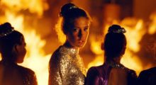 Women wearing long-sleeve, backless, sequin gowns stand in front of a roaring fire. One turns to face the camera.