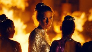 Women wearing long-sleeve, backless, sequin gowns stand in front of a roaring fire. One turns to face the camera.
