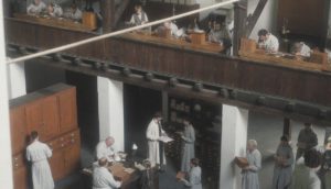 Monks in long, white robes work on watches in a multi-level building embellished with dark wood fixtures.