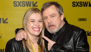 Claudette Godfrey, a young woman with straight, blond hair and red lipstick, poses with actor Mark Hamill, who has salt and pepper hair and a beard.