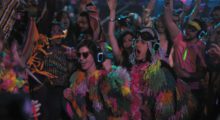 A gaggle of partygoers wear fringed rainbow jackets and ornate accessories as they rave.
