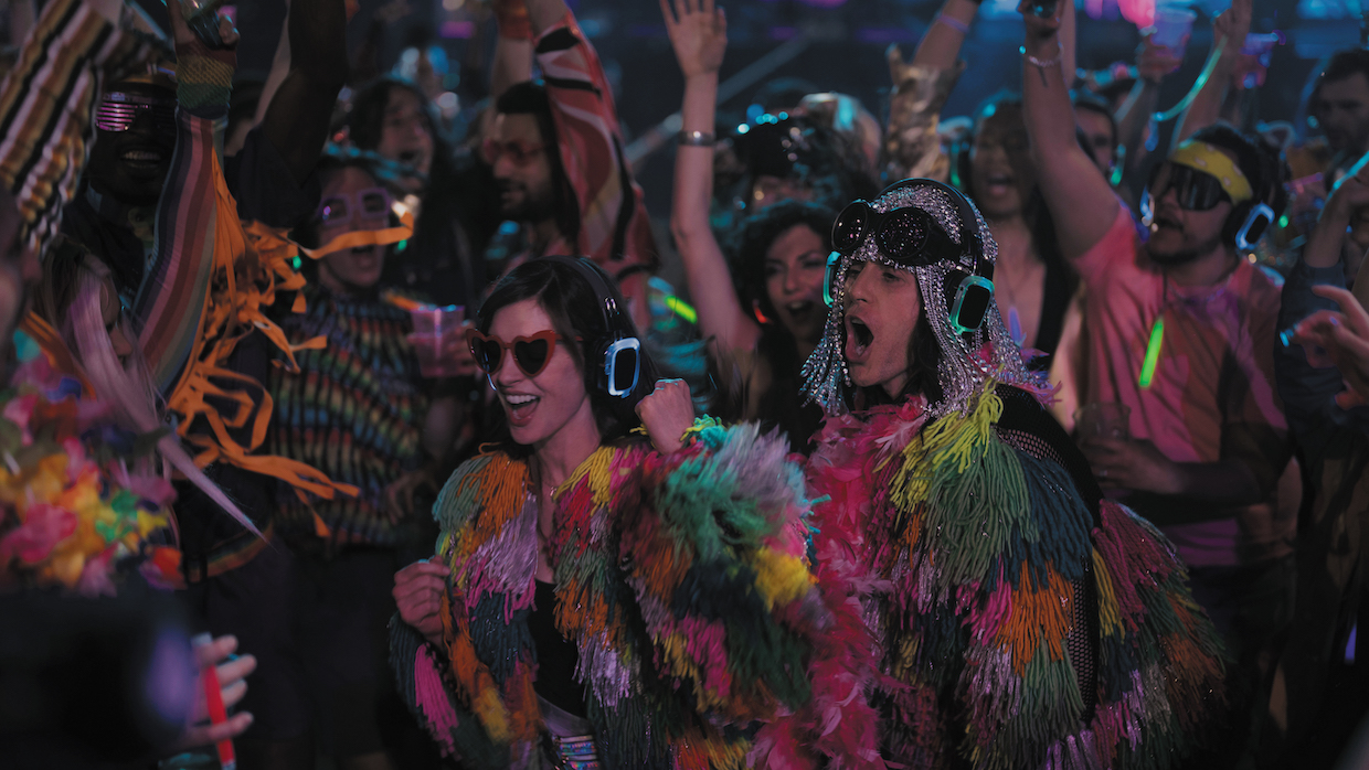 A gaggle of partygoers wear fringed rainbow jackets and ornate accessories as they rave.