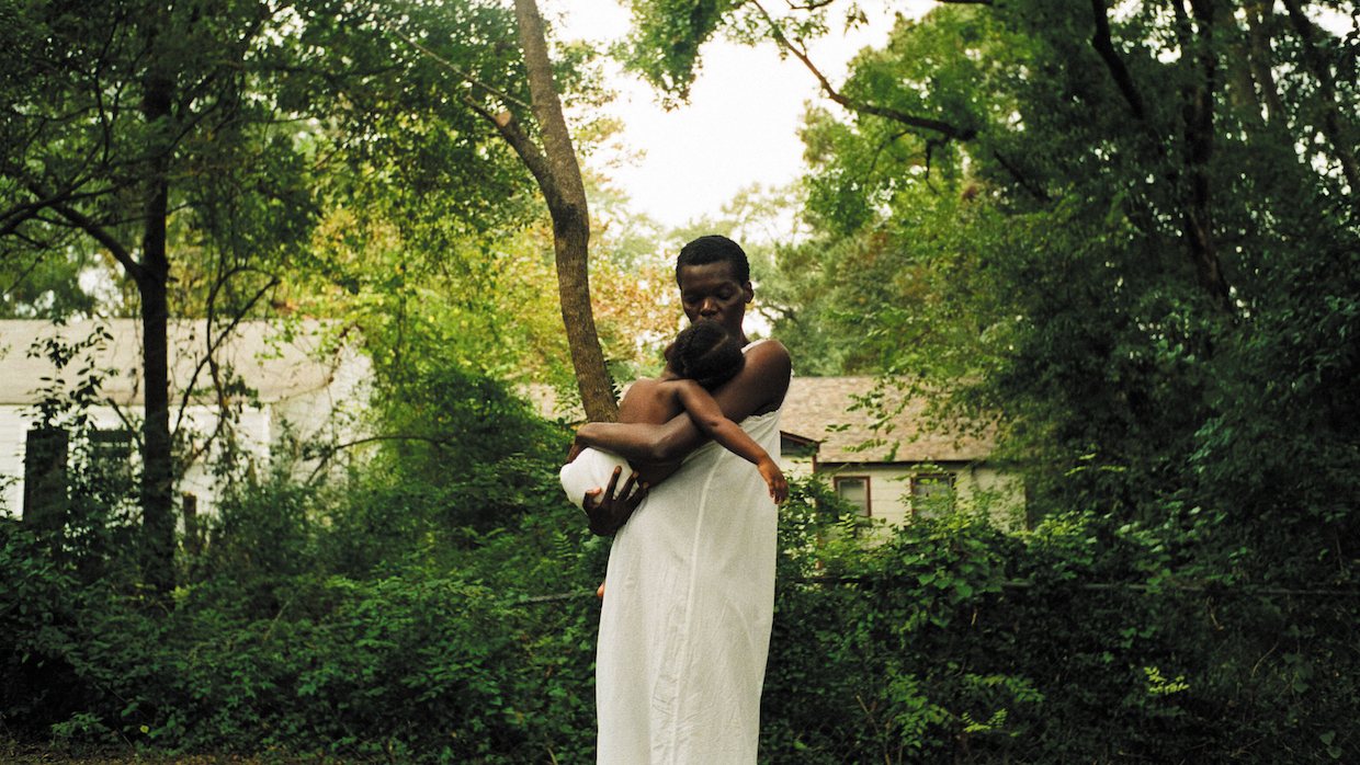 A person wearing an off-white long tunic holds a baby in their arms. They are surrounded by lush, green trees and a semi-obscured house.