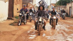 A group of young people ride their motorcycles through damp streets in a Brazilian town.