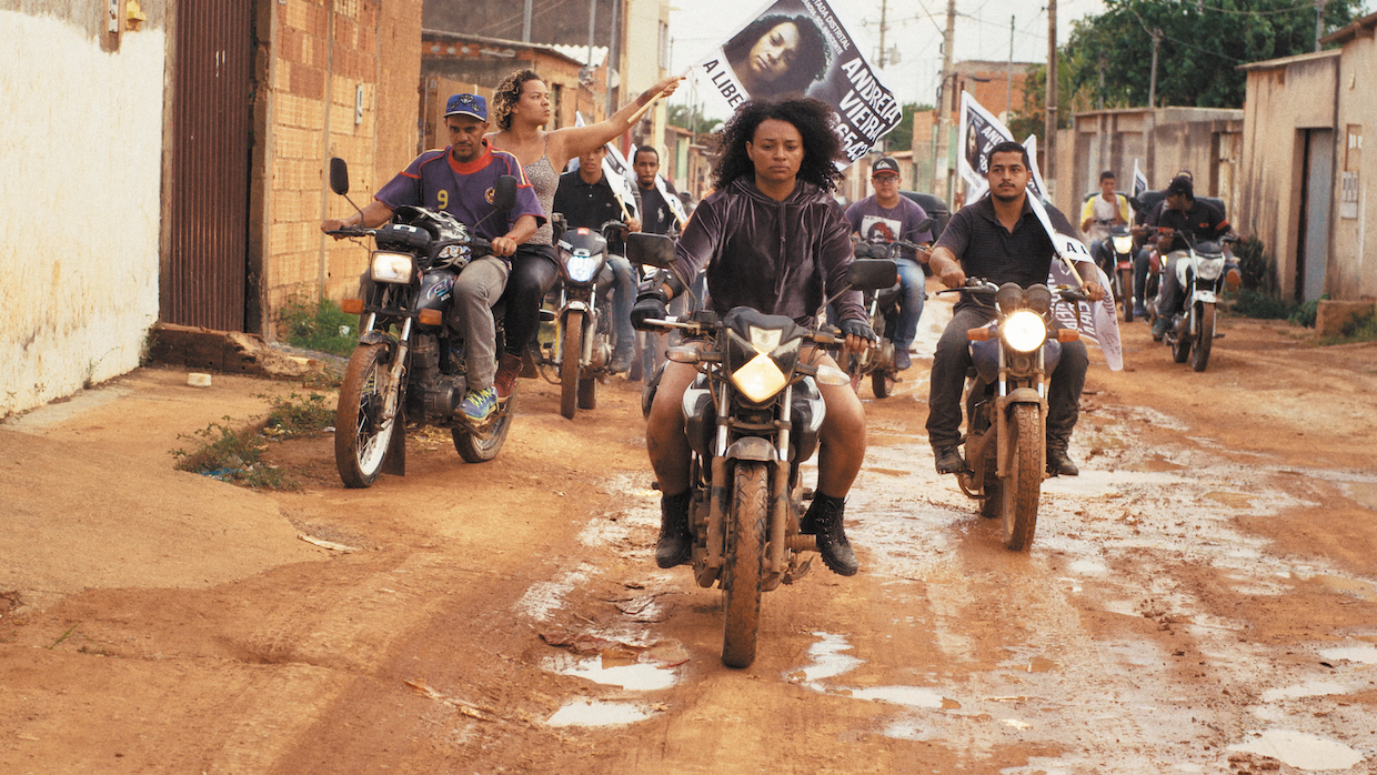 A group of young people ride their motorcycles through damp streets in a Brazilian town.