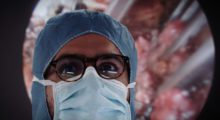 A doctor wearing surgical scrubs stands before an enlarged image of internal organs.