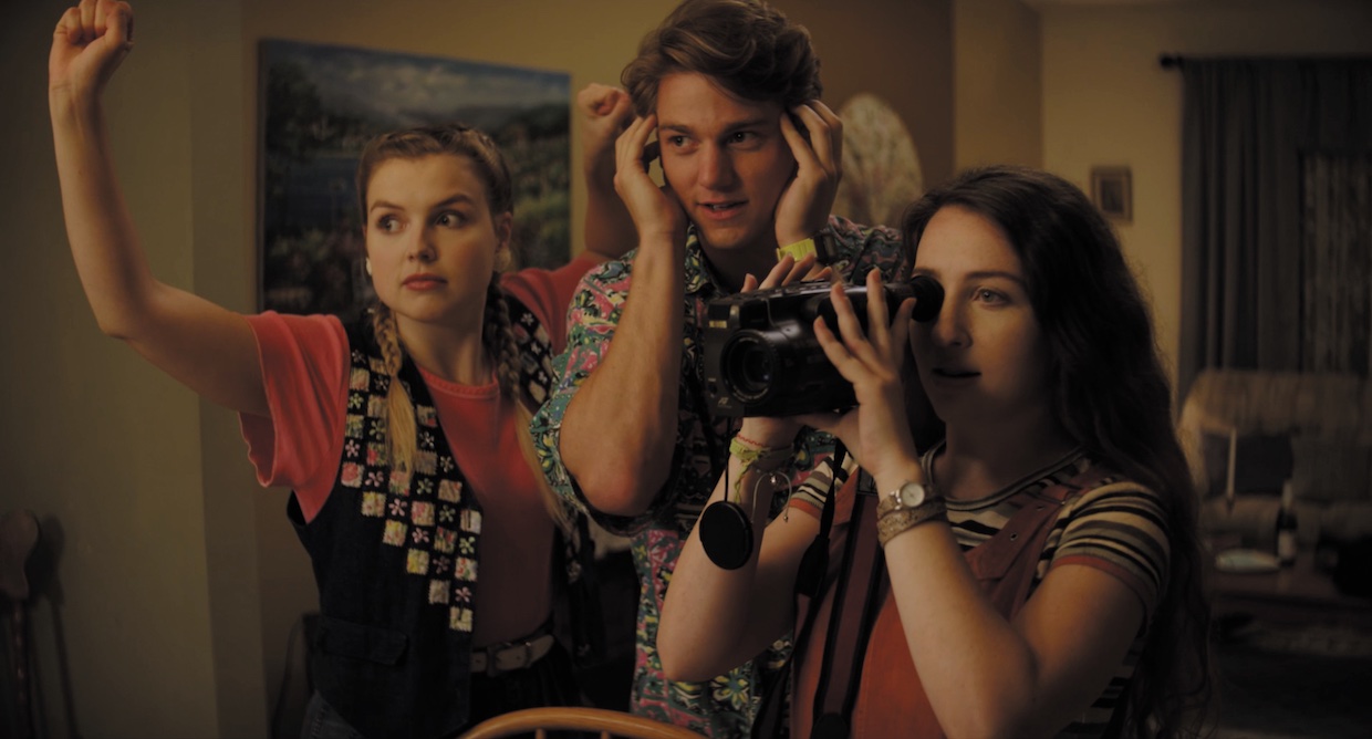 Three young people wield (oft-imaginary) moviemaking objects: A girl with blond hair pretends to hoist a boom mic, a boy with brown hair pretends to listen to audio on invisible headphones and a short girl with brown hair holds a camcorder.