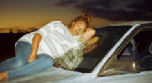 A pregnant woman wearing a white and gray striped t-shirt and blue jeans lounges atop the hood of a car. Her brown hair is tied up in a messy bun.