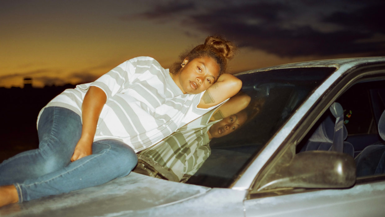 A pregnant woman wearing a white and gray striped t-shirt and blue jeans lounges atop the hood of a car. Her brown hair is tied up in a messy bun.